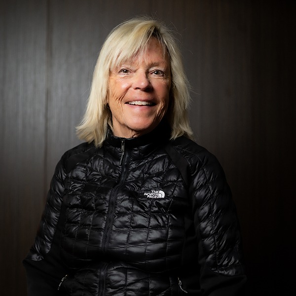 Smiling blonde woman wearing a black puffer jacket, standing against a dark wood background. - Breck Film | Eclipse Theater