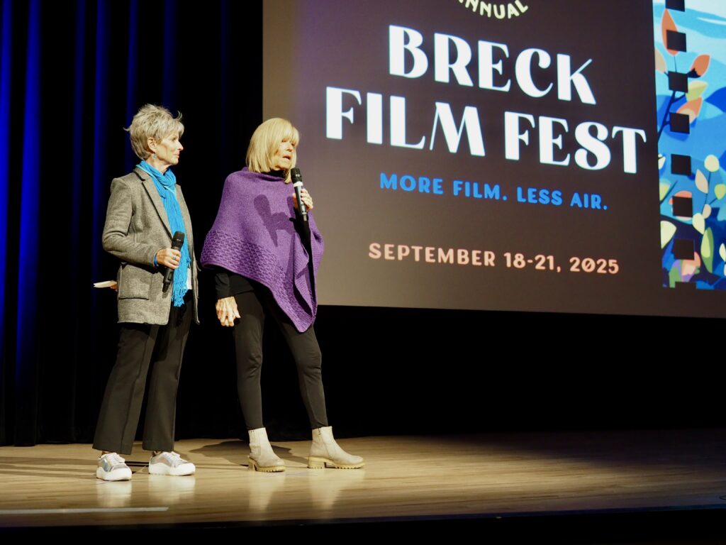 OLYMPUS DIGITAL CAMERA - Breck Film | Eclipse Theater Two women stand on a stage holding microphones, with a large Breck Film Fest banner on screen behind them. - Breck Film | Eclipse Theater