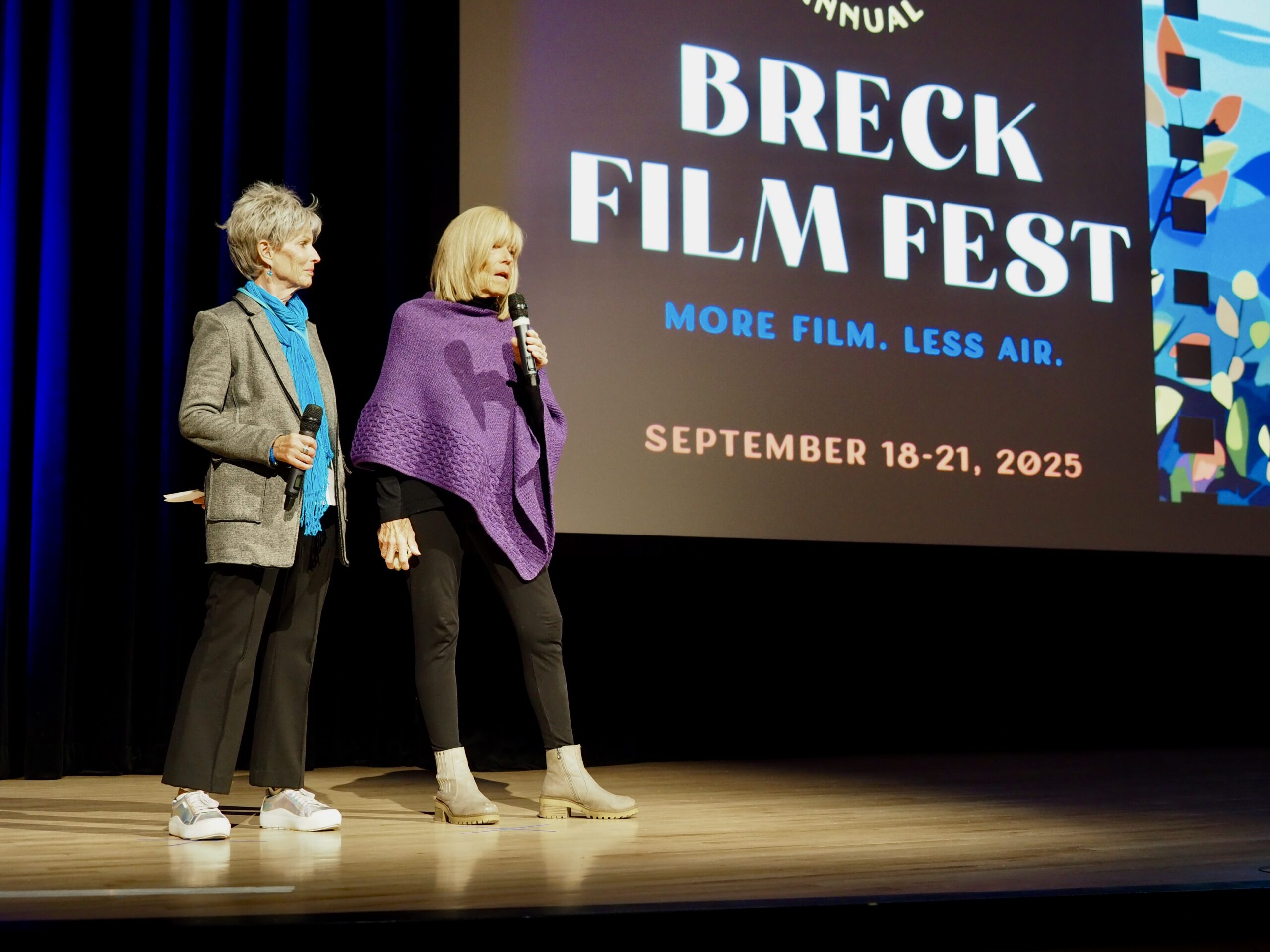 Two women stand on a stage holding microphones, with a large Breck Film Fest banner on screen behind them. - Breck Film | Eclipse Theater