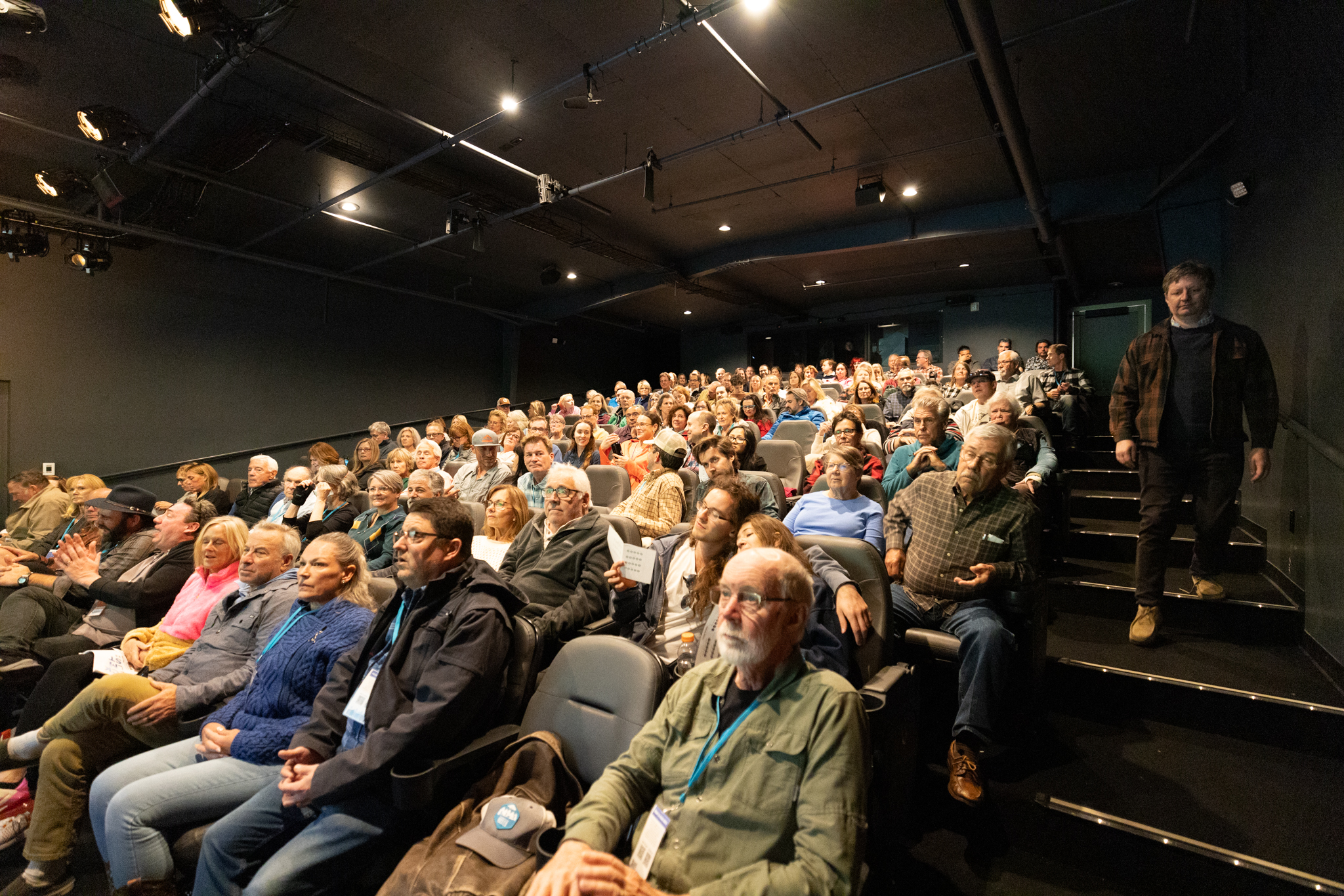 Large audience seated in a dim theater, looking toward the stage; a man in a brown jacket stands on the right by the exit stairs.”] , - Breck Film | Eclipse Theater